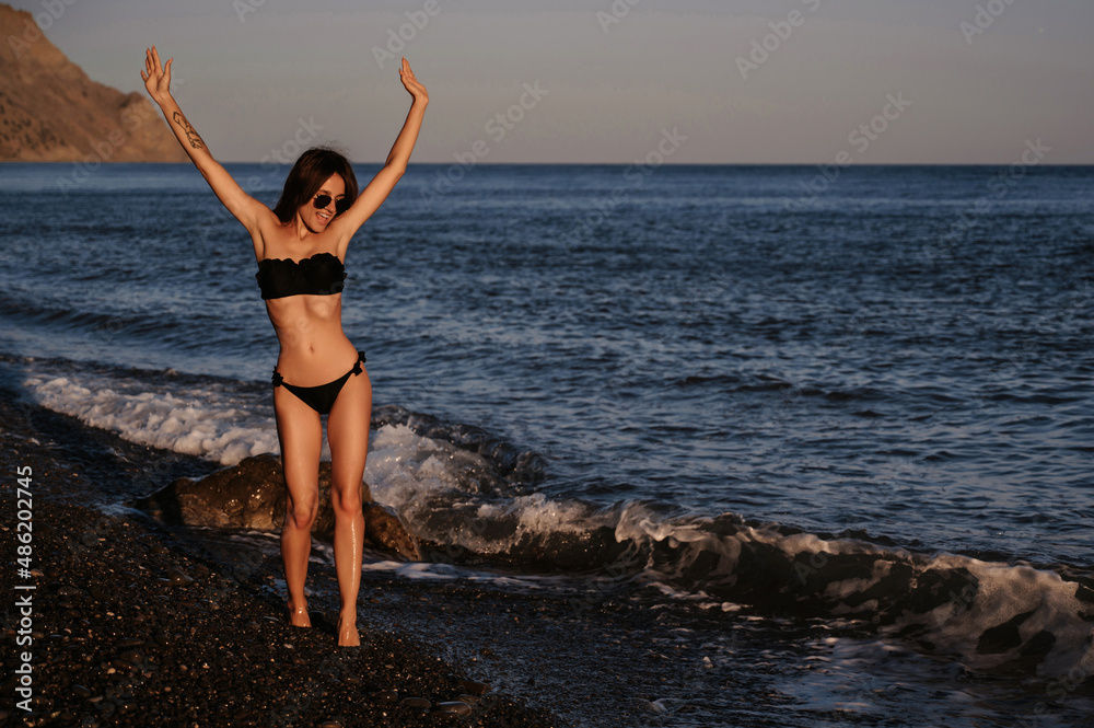 young girl with a sexy body in a black swimsuit and sunglasses enjoys relaxing on the beach by the sea in summer