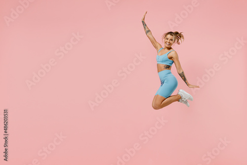 Full body young strong sporty athletic fitness trainer instructor woman wearing blue tracksuit spend time in home gym jump high stretch hands isolated on plain pink background. Workout sport concept.