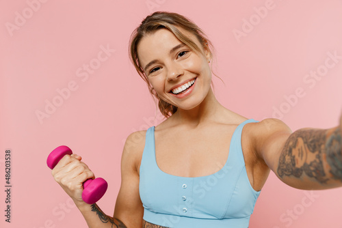Young strong fitness trainer instructor woman wear blue tracksuit spend time in home gym hold female dumbbells do selfie shot on mobile phone isolated on plain pink background. Workout sport concept