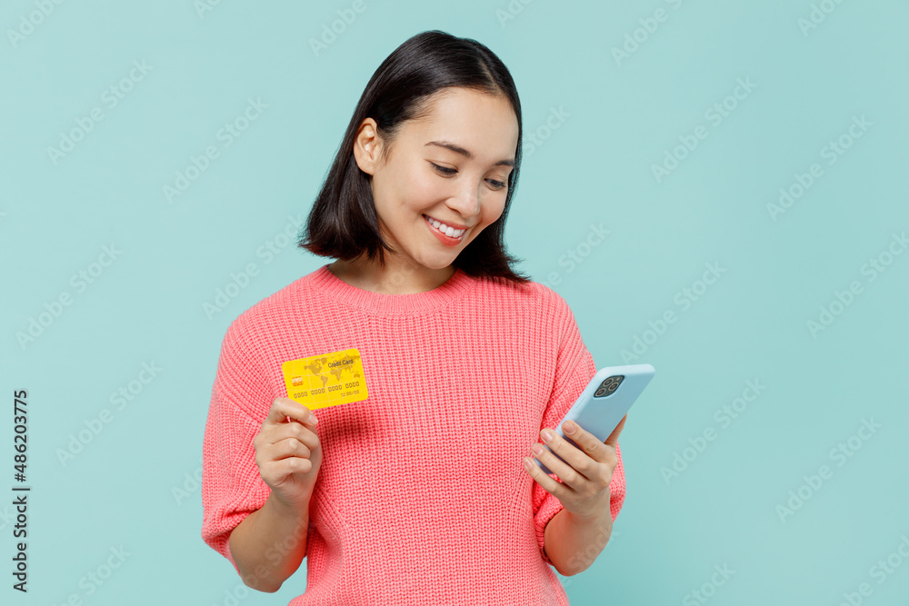 Young woman of Asian ethnicity 20s wearing pink sweater using mobile cell phone hold credit bank card doing online shopping order delivery booking tour isolated on pastel plain light blue background. Young woman of Asian ethnicity 20s wearing pink sweater using mobile cell phone hold credit bank card doing online shopping order delivery booking tour isolated on pastel plain light blue background.