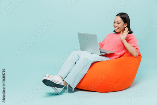 Full body young smiling happy woman of Asian ethnicity 20s wear pink sweater sit in bag chair talk by video call on laptop pc computer waving hand isolated on pastel plain light blue color background. photo