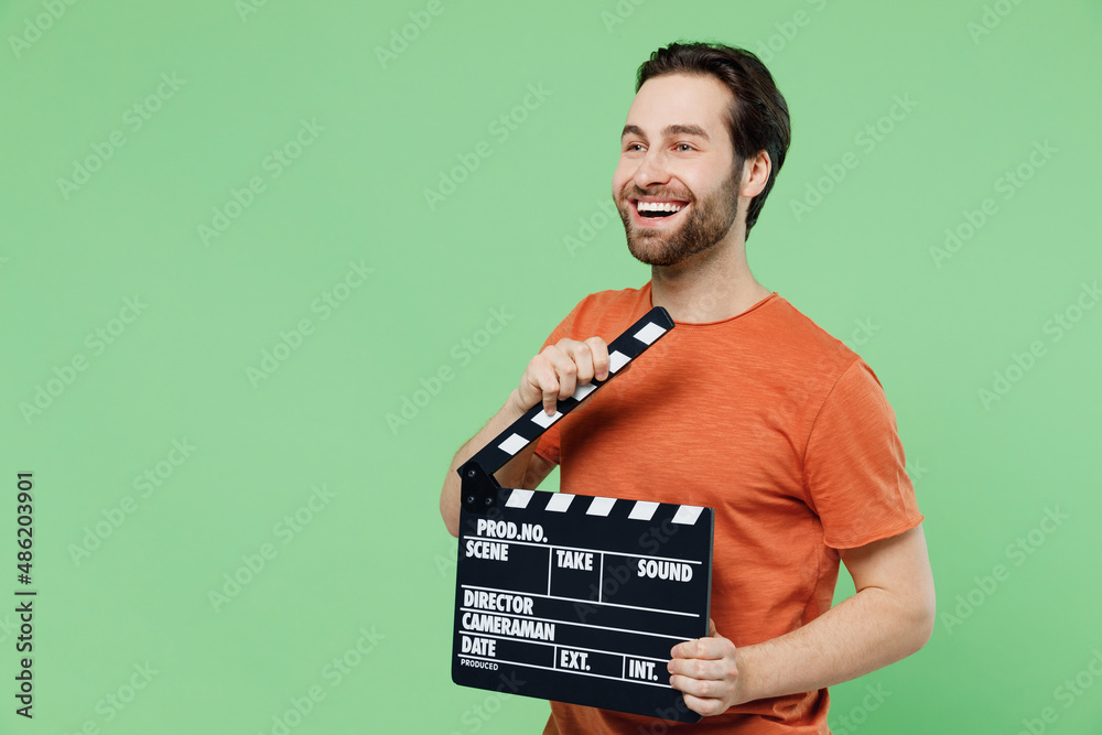 Young smiling happy man 20s in casual orange t-shirt holding classic black film making clapperboard look aside on workspace area isolated on plain pastel light green color background studio portrait