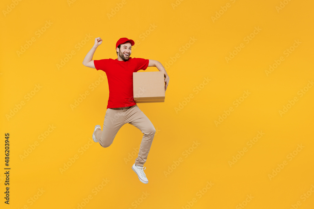 Full body fun happy delivery guy employee man in red cap T-shirt uniform workwear work as dealer courier jump high hold cardboard box do winner gesture run isolated on plain yellow background studio.
