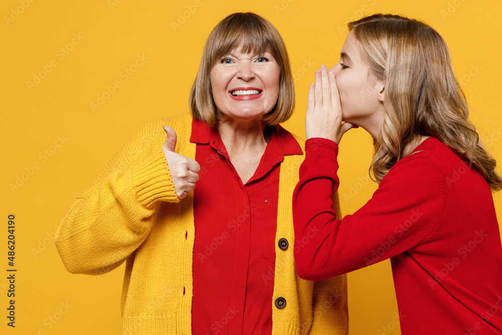 Woman 50s in red shirt have fun with teenager girl 12-13 years old. Grandmother granddaughter whisper gossip tells secret behind her hand share news show thumb up isolated on plain yellow background Woman 50s in red shirt have fun with teenager girl 12-13 years old. Grandmother granddaughter whisper gossip tells secret behind her hand share news show thumb up isolated on plain yellow background