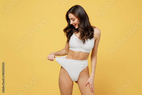 Happy fun satisfied lovely attractive young brunette woman 20s in white underwear with perfect fit body look aside show losing weight result hold panties isolated on plain yellow background studio