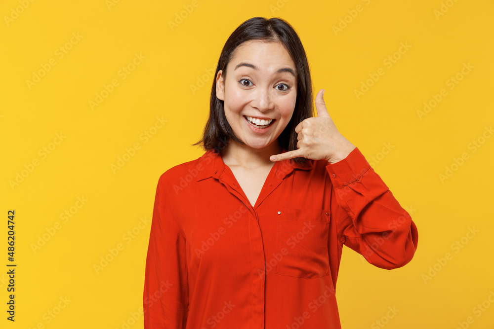Smiling stunning cheerful young woman of Asian ethnicity 20s years old wears orange shirt looking camera doing phone gesture like says call me back isolated on plain yellow background studio portrait. Smiling stunning cheerful young woman of Asian ethnicity 20s years old wears orange shirt looking camera doing phone gesture like says call me back isolated on plain yellow background studio portrait.