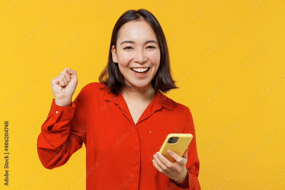 Jubilant vivid excited fancy young woman of Asian ethnicity 20s years old wear orange shirt hold in hand use mobile cell phone doing winner gesture isolated on plain yellow background studio portrait Jubilant vivid excited fancy young woman of Asian ethnicity 20s years old wear orange shirt hold in hand use mobile cell phone doing winner gesture isolated on plain yellow background studio portrait