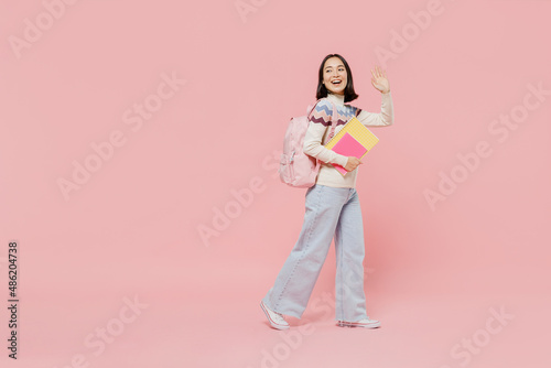 Full body side view teen fun student girl of Asian ethnicity wearing sweater hold backpack book walk going waving hand isolated on pastel plain pink background Education in university college concept