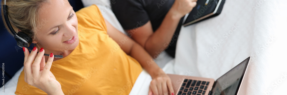 © megaflopp - Young woman in headphones with microphone works on laptop while lying in bed