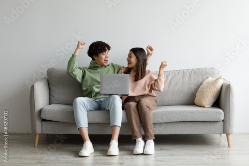 Overjoyed young Asian couple with laptop gesturing YES, celebrating good news or success, sitting on sofa at home