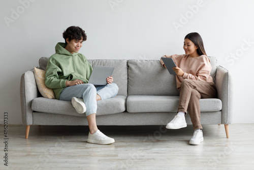 Handsome young Asian guy and his girlfriend using modern gadgets while sitting on couch at home, copy space