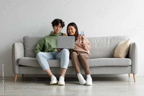 Online shopping. Young Asian couple with laptop and credit card sitting on couch, buying goods in web store at home