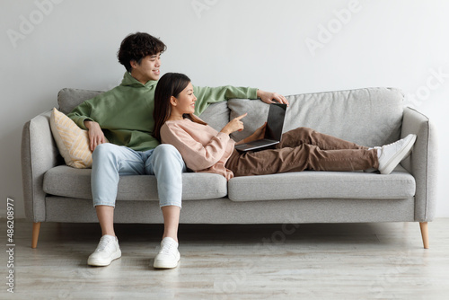Loving young Asian couple cuddling together on couch with laptop, watching movie together, having online video chat