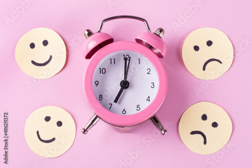 Mechanical classic alarm clock (hands at 7 o'clock in the morning), funny and sad emoticons on a pink background. The concept of early awakening (wake up). Good and bad moods in the morning.