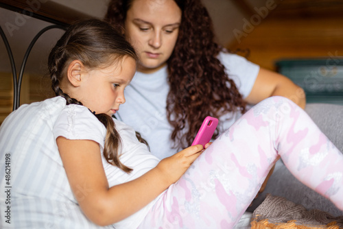 Serious young girl holding phone in hands with mother lying on bed both looking at phone in wooden house wearing home clothes in daytime. Family time together.