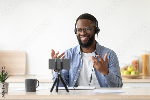 Friendly glad young african american bearded guy teacher in glasses and headphones shooting video lesson for vlog photo