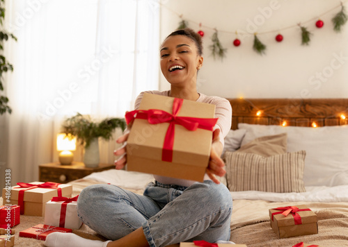 Overjoyed african american lady holding and giving wrapped present box to camera, resting in decorated bedroom © Prostock-studio