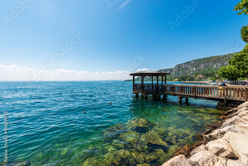Fototapeta Naklejka Na Ścianę i Meble -  Lake Garda in front of the small Garda town. Beautiful bay with a wooden pier with roof and the headland of San Vigilio (Punta San Vigilio). Tourist resort in Verona province, Veneto, Italy, Europe.