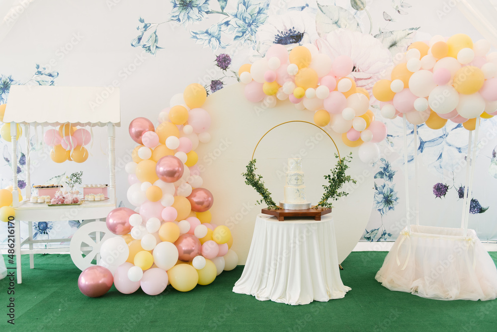 A large three-tiered white cake for the baptism of a child with green twigs on the background of a beautiful photo zone with balloons and a candy bar