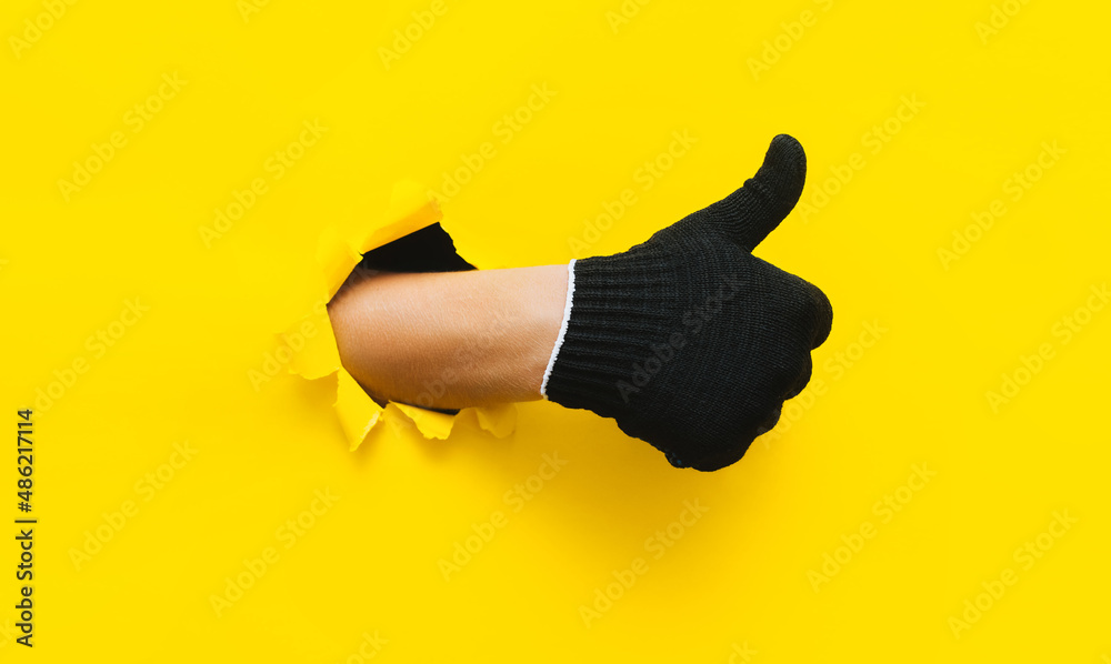 A right man's hand in a black fabric work glove shows a thumb up (like). Torn hole in yellow paper. Good job, fun mood and approval concept. Copy space.