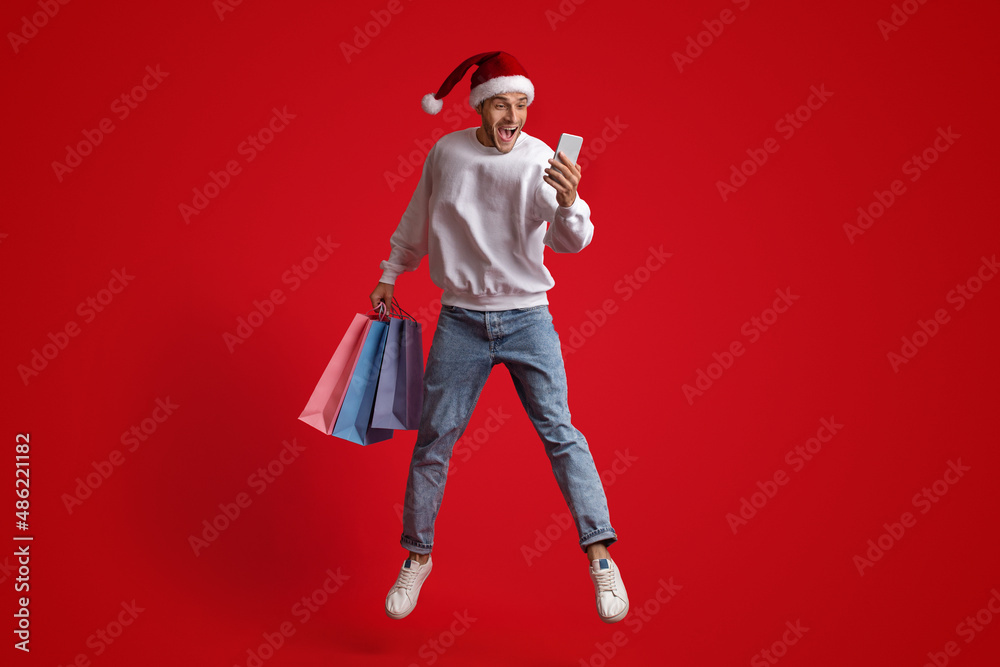 Excited Guy In Santa Hat Jumping With Smartphone And Shopping Bags Excited Guy In Santa Hat Jumping With Smartphone And Shopping Bags