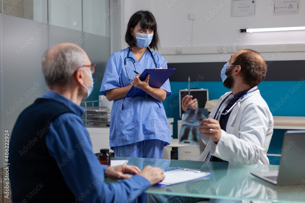 Obraz premium Physician talking to nurse and holding x ray scan at checkup visit with retired man in office. Doctor explaining radiography diagnosis to patient while he asks medical assistant to help.