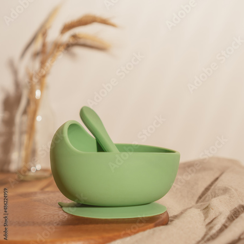 Modern green silicone bowl with suction base and spoon on wooden table near glass vase with dried flowers. Serving food, baby tableware, first feeding concept. Instagram use, square frame, soft focus.