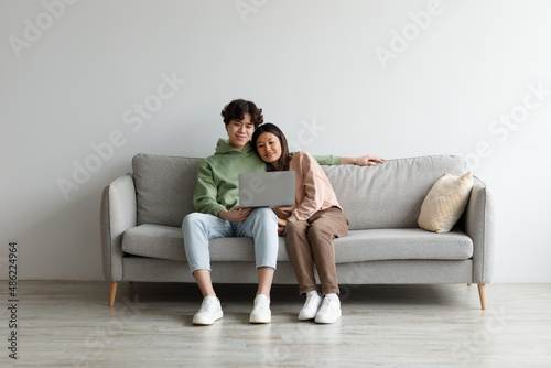 Affectionate millennial Asian couple using laptop computer, chilling together on couch at home, free space © Prostock-studio