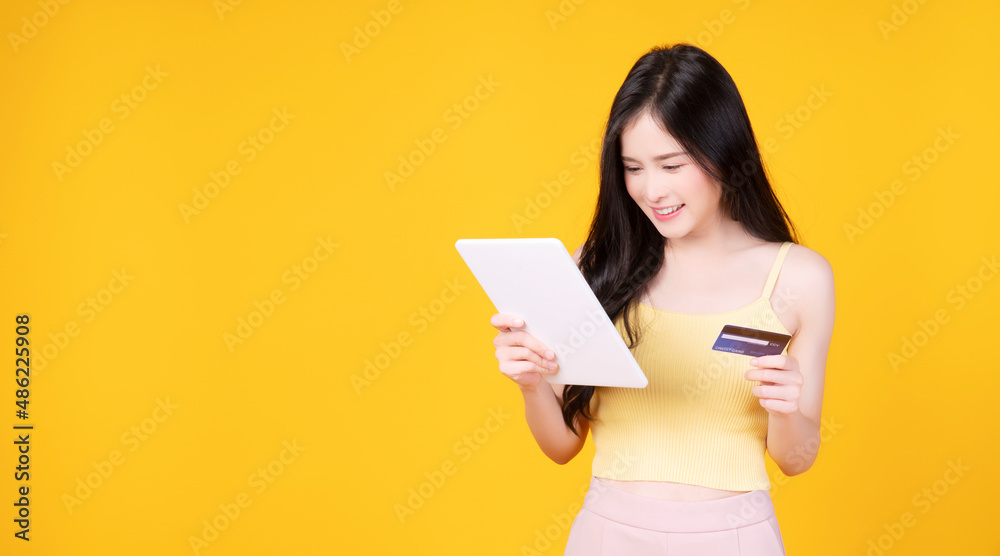 Joyful Asian woman holding credit card looking at tablet on yellow background. Pretty young girl using digital tablet surfing shopping online paying money credit card over isolated. Technology banking Joyful Asian woman holding credit card looking at tablet on yellow background. Pretty young girl using digital tablet surfing shopping online paying money credit card over isolated. Technology banking