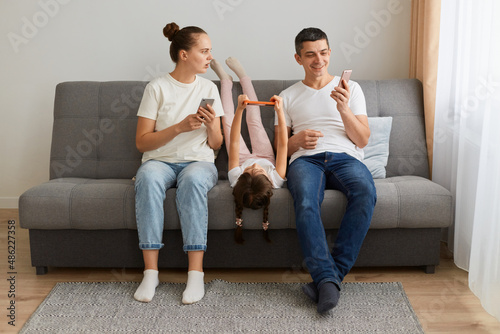 Indoor shot of family sitting on sofa together father showing shock content and laughing, woman looking with open mouth and surprised expression, gadget addiction.
