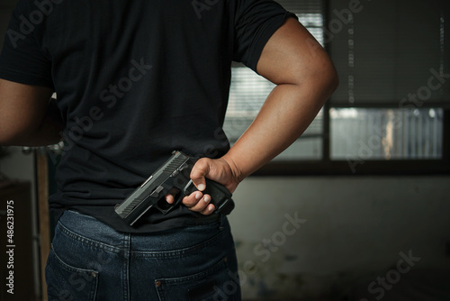 man holding a pistol behind his back standing in the room in black concept of assassination, murder, criminal © Tanawit