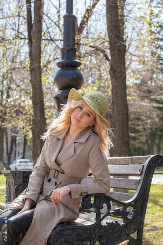 Beautiful blonde woman in a raincoat and hat sits on a bench