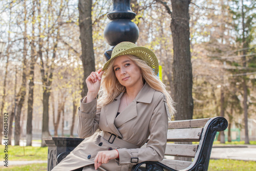 Beautiful blonde woman in a raincoat and hat sits on a bench
