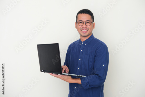 Adult Asian man looking to the camera while holding a laptop and showing happy expression