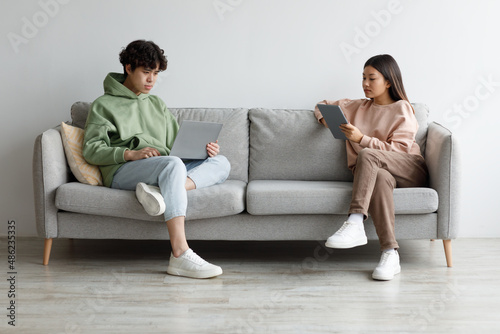 Serious young Asian woman and her boyfriend using laptop and tablet computers, browsing internet on couch at home