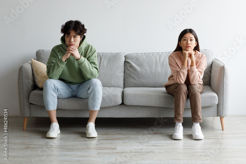 Stubborn young Asian couple sitting on opposite sides of couch in silence after fight, indoors
