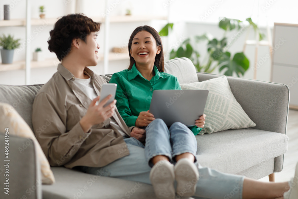 Married Asian couple using laptop and smartphone at home, relaxing on couch together, watching movies, chatting online Married Asian couple using laptop and smartphone at home, relaxing on couch together, watching movies, chatting online