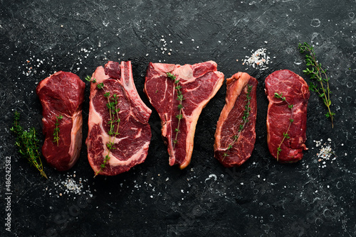 A variety of raw beef steaks for grilling with seasonings: T-bone, striploin steak, ribeye. Top view. On a black stone background.