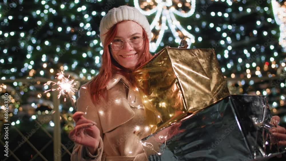 festive atmosphere, portrait of smiling woman in hat and glasses with gifts in hand and with bingal lights stands backdrop of Christmas tree and lights at night in park festive atmosphere, portrait of smiling woman in hat and glasses with gifts in hand and with bingal lights stands backdrop of Christmas tree and lights at night in park