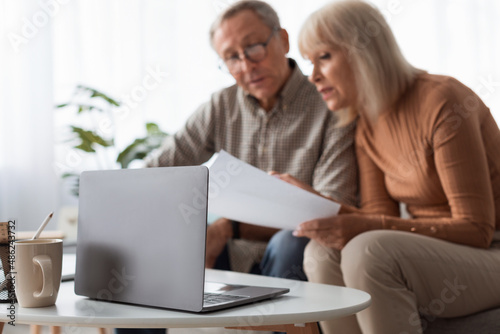 Senior Couple Reading Papers Using Laptop Sitting On Couch Indoors