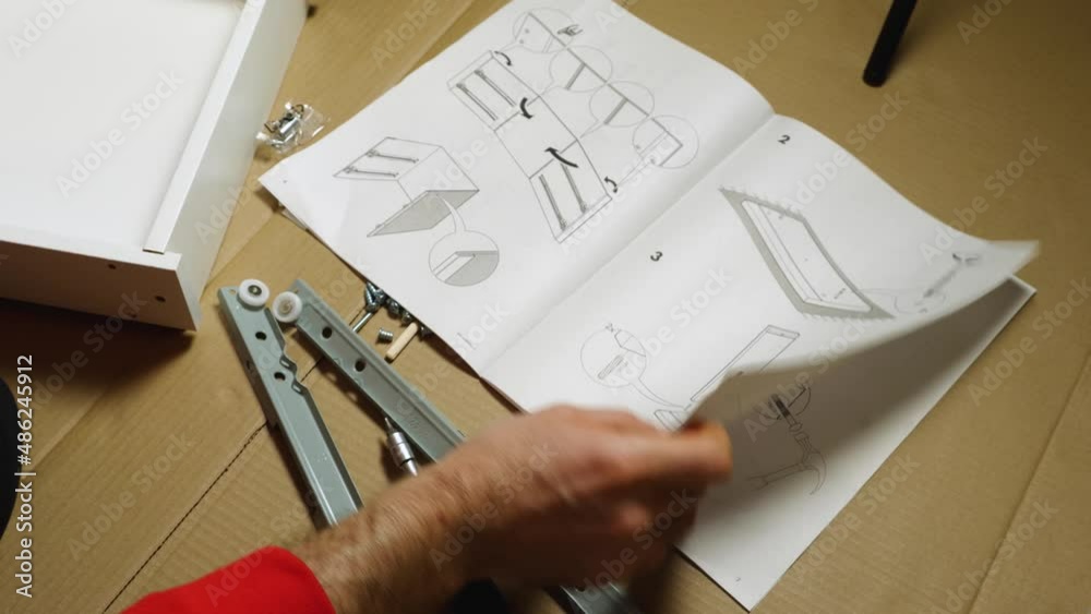 Close-up of a man's hand leafing through instructions for assembling furniture. Nearby on cardboard are tools, screws and furniture elements. Assembling furniture at home with your own hands