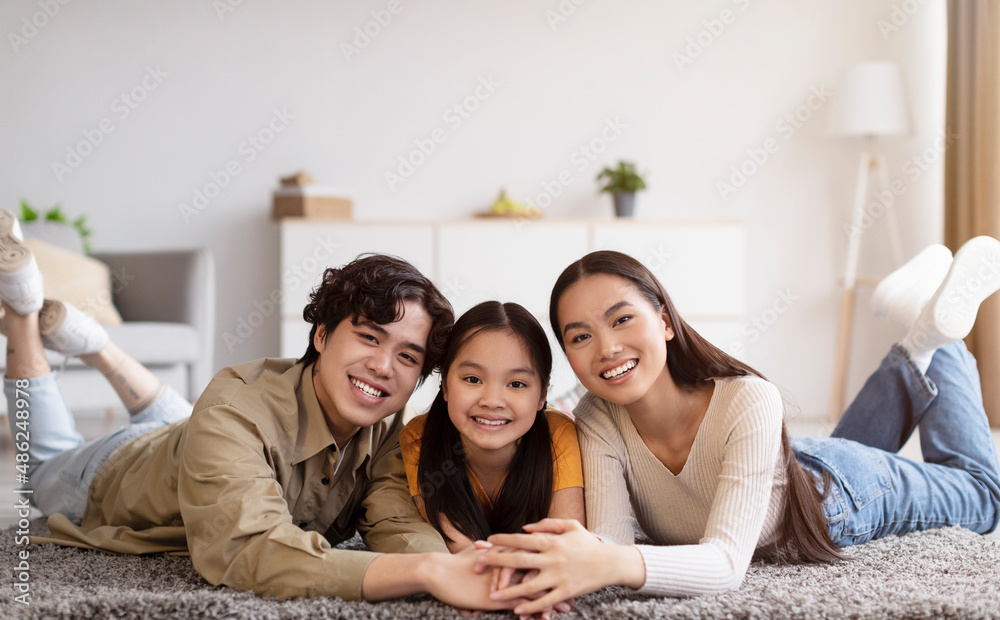 Cheerful millennial asian lady, guy and teen daughter lie on floor and hold hands in living room interior Cheerful millennial asian lady, guy and teen daughter lie on floor and hold hands in living room interior