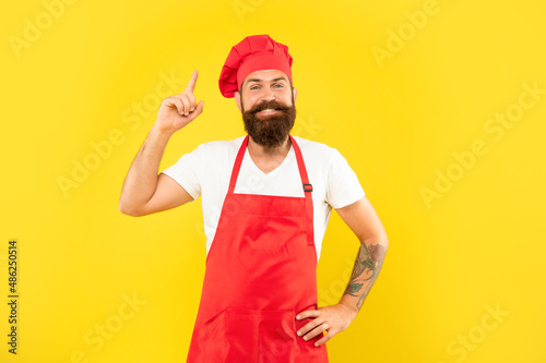 Happy man cook in toque and apron keeping finger raised yellow background  idea