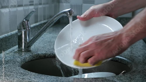 Close-up young man washing dishes at home with soap and sponge. The guy washes dishes every day. Using too much water. The concept of unsustainable resource consumption