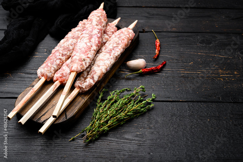 Raw Lula kebab on skewers, with grill ingredients, on black wooden table background, with copy space for text