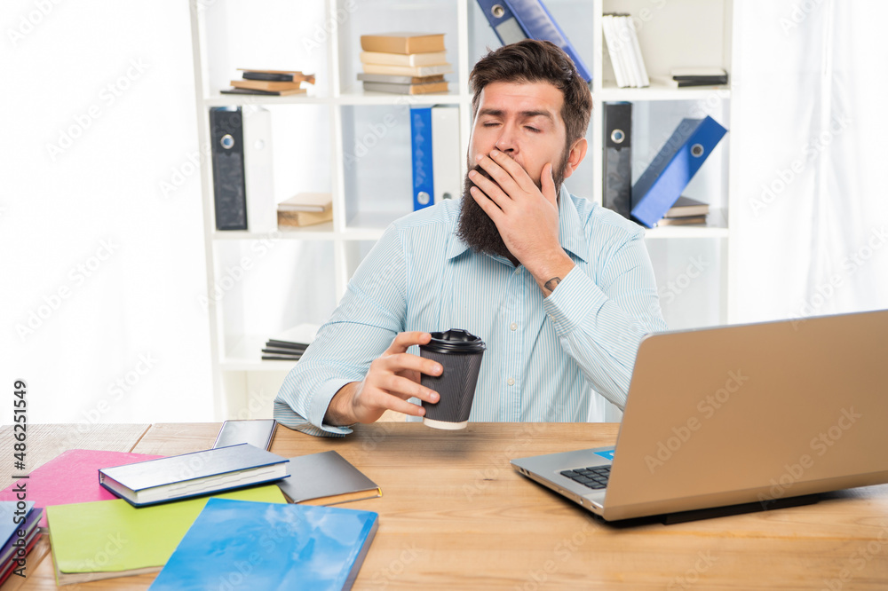 Sleepy businessman yawning and drinking takeaway coffee at office desk, morning