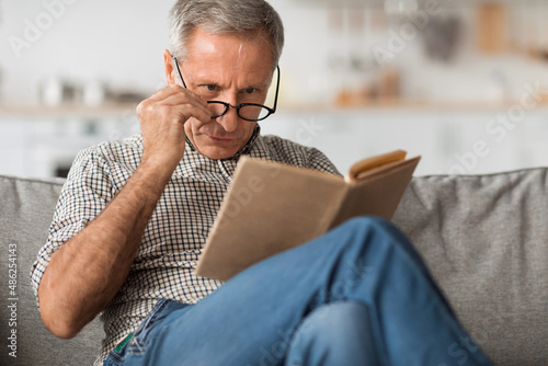 Senior Man With Poor Eyesight Reading Book Above Eyeglasses Indoor photo