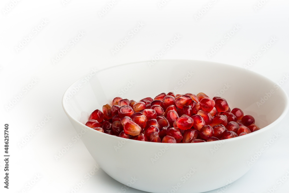 Red pomegranate seeds in a white plate, on the white background. Fresh juicy pomegranate seeds isolated on white. Close-up, side view, selective focus, copy space Red pomegranate seeds in a white plate, on the white background. Fresh juicy pomegranate seeds isolated on white. Close-up, side view, selective focus, copy space