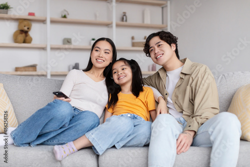 Smiling millennial korean mother, father and teen girl hugging and watch tv, sit on sofa in living room