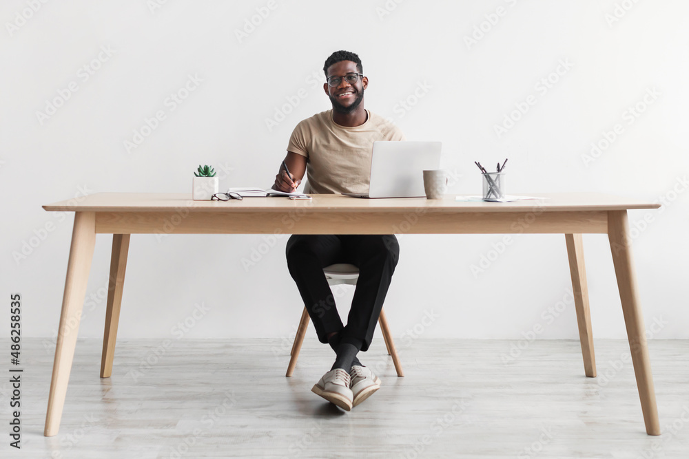 Smiling African American man writing in notebook, taking notes and using computer, sitting at desk in home office, copy space